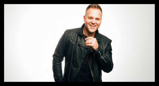 Matthew West Smiling While Lifting His Hand In Front Of A White Background